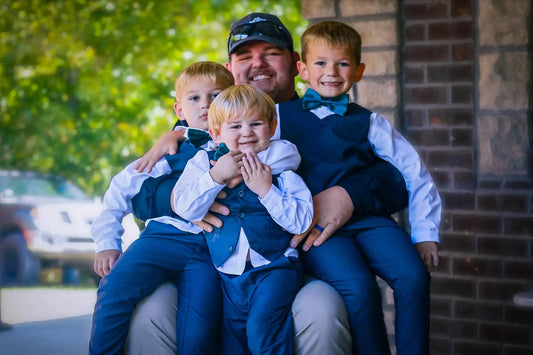 Father with three young boys in matching blue suits outdoors, smiling for a family wedding portrait.