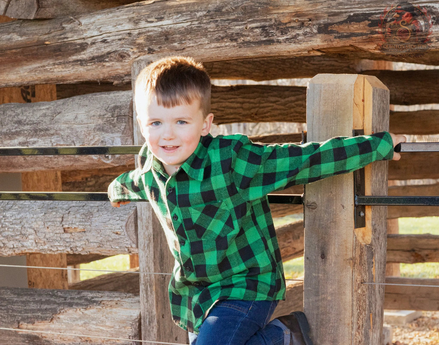 Young boy in green plaid shirt smiling, leaning on a rustic log fence outdoors, Quinton Stocton Photography.