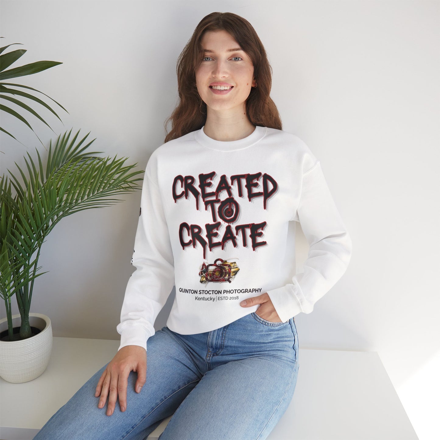 Woman wearing a white sweatshirt with text and graphics, sitting on a white surface with a plant in the background.