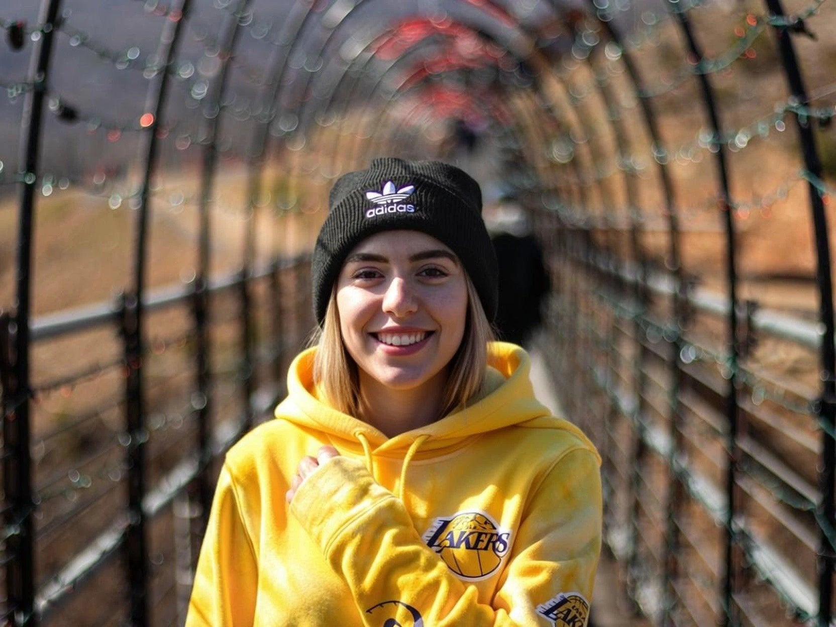 Smiling woman in yellow Lakers hoodie and black Adidas beanie on a sunlit tunnel bridge