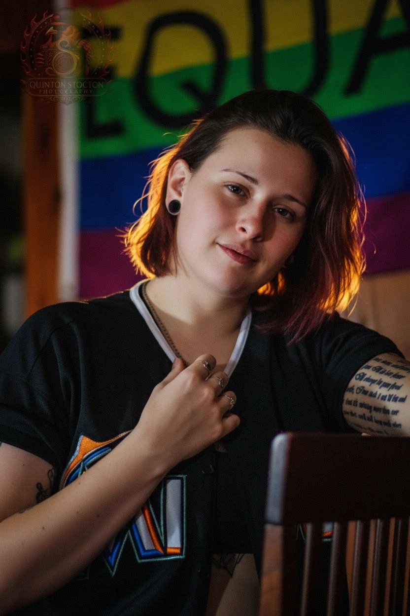 Portrait of a woman with short hair and tattoos in a black tee, warm backlight, rainbow backdrop.