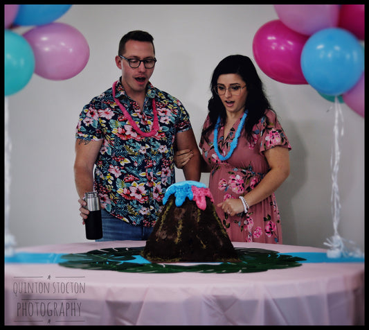 Event photography showing a couple revealing baby's gender with a volcano cake erupting blue & pink foam. Baby gender reveal